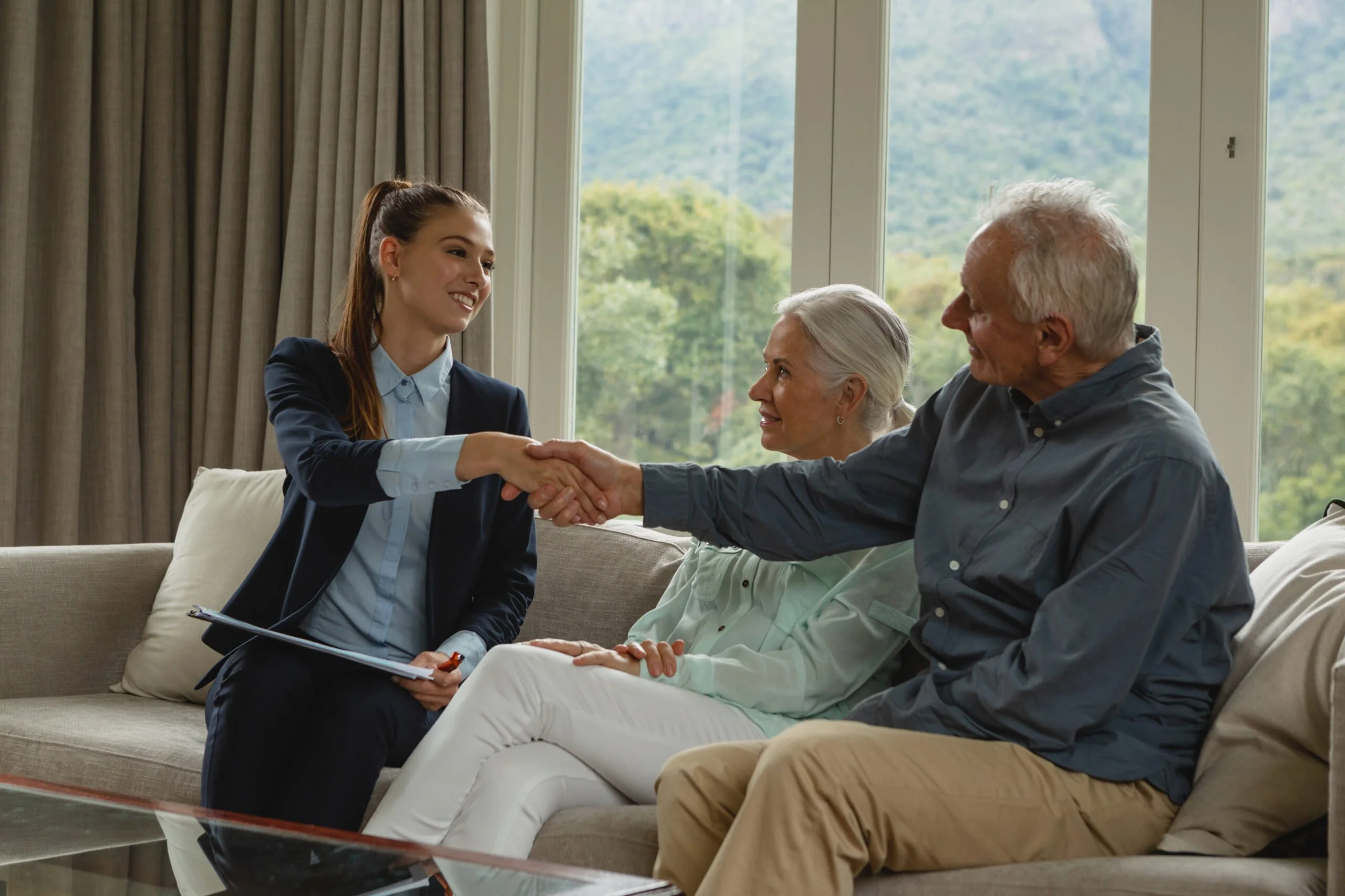 Senior man sitting on a sofa shaking hands with a real estate agent while a senior woman looks on in a living room.
